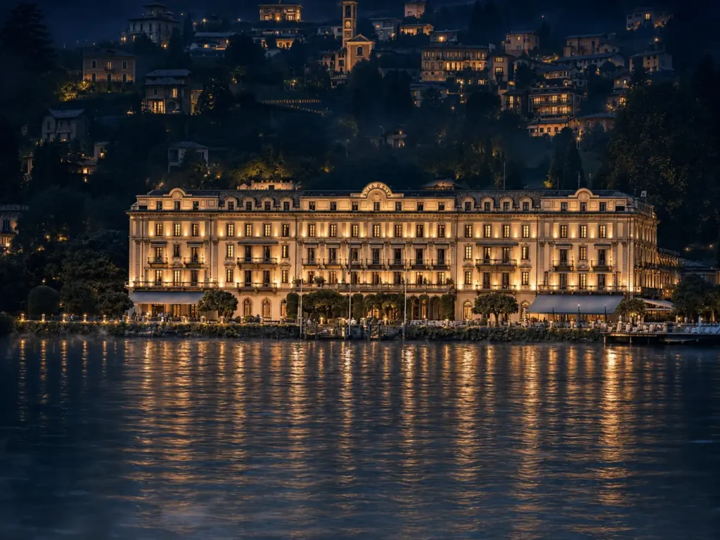 Un grand hôtel illuminé au bord d’un lac, avec ses reflets sur l’eau et un village sur la colline à l’arrière-plan.