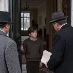 Un garçon ouvre la porte à deux hommes coiffés de chapeaux, dont l’un porte une serviette et des documents.
